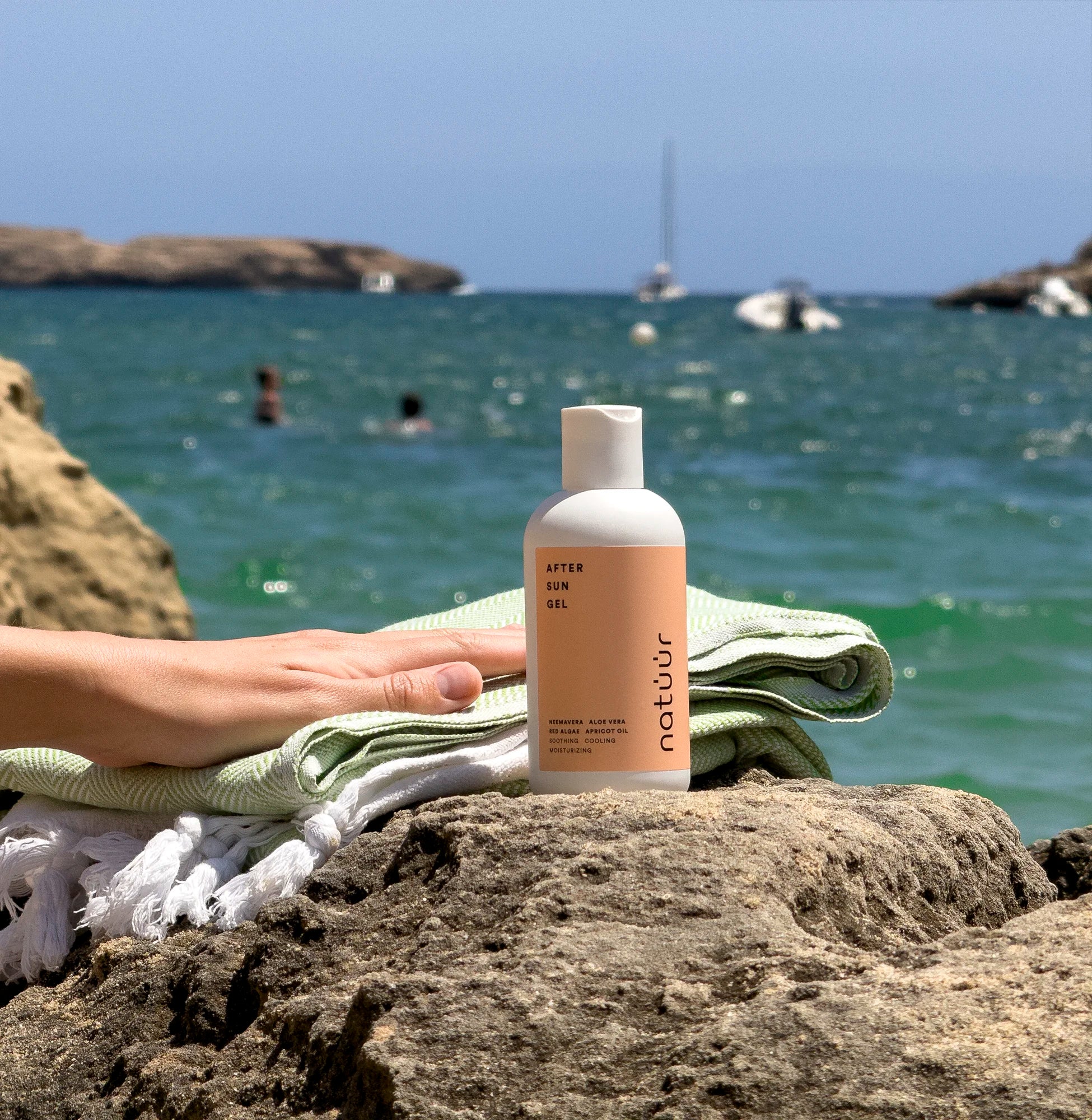Man sieht das After Sun Gel der Marke natüür auf einem Stein, im Hintergrund das blaue Meer. Das Label auf der weißen Plastikflasche ist pfirsichfarben. Neben dem Produkt liegt ein Handtuch und man sieht eine Hand, die danach greift.