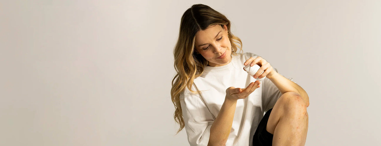 Eine Frau mit blond-braunen Haaren und weißem T-Shirt sitzt auf dem Boden. Ihr Knie ist angewinkelt und von Psoriasis betroffen. In der Hand hält sie die Pflegecreme von natüür.