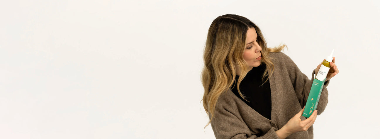 Eine Frau mit langen braunen Haaren und einer braunen Jacke hat die grüne Umverpackung des Scalp Tonics der Marke natüür in der Hand. Mit der anderen Hand zieht sie die Flasche mit dem Scalp Tonic raus.