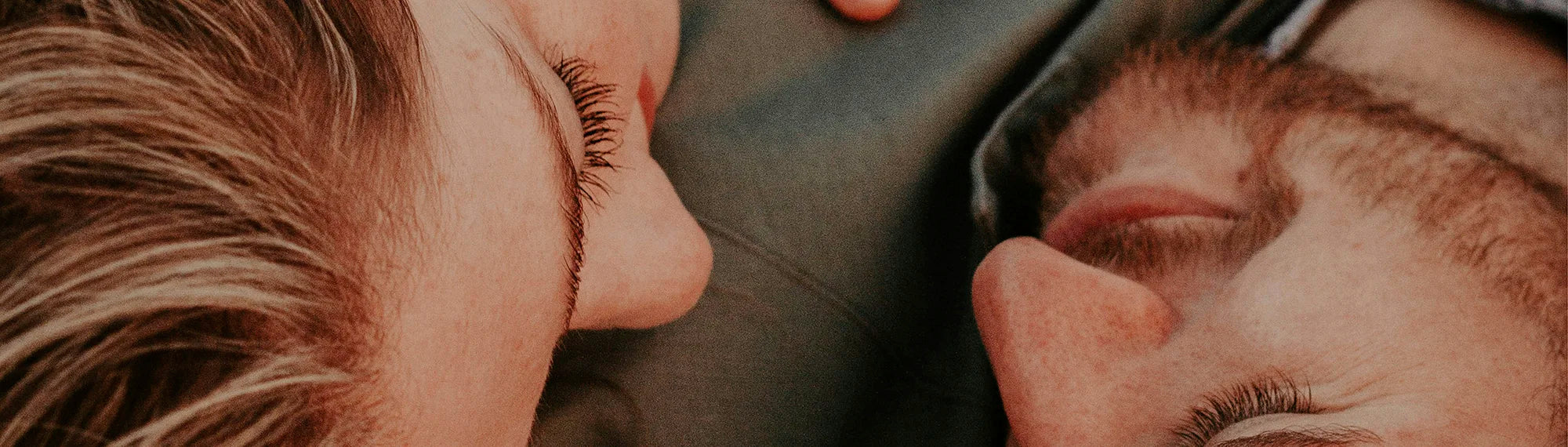 Ein Mann mit dunklem Bart und eine Frau mit blonden Haaren sind liegend zu sehen in einer Nahaufnahme. Sie hat ihren Kopf auf seiner Brust und die Hand auf seinem Oberkörper. Sie schauen sich an und wirken glücklich.