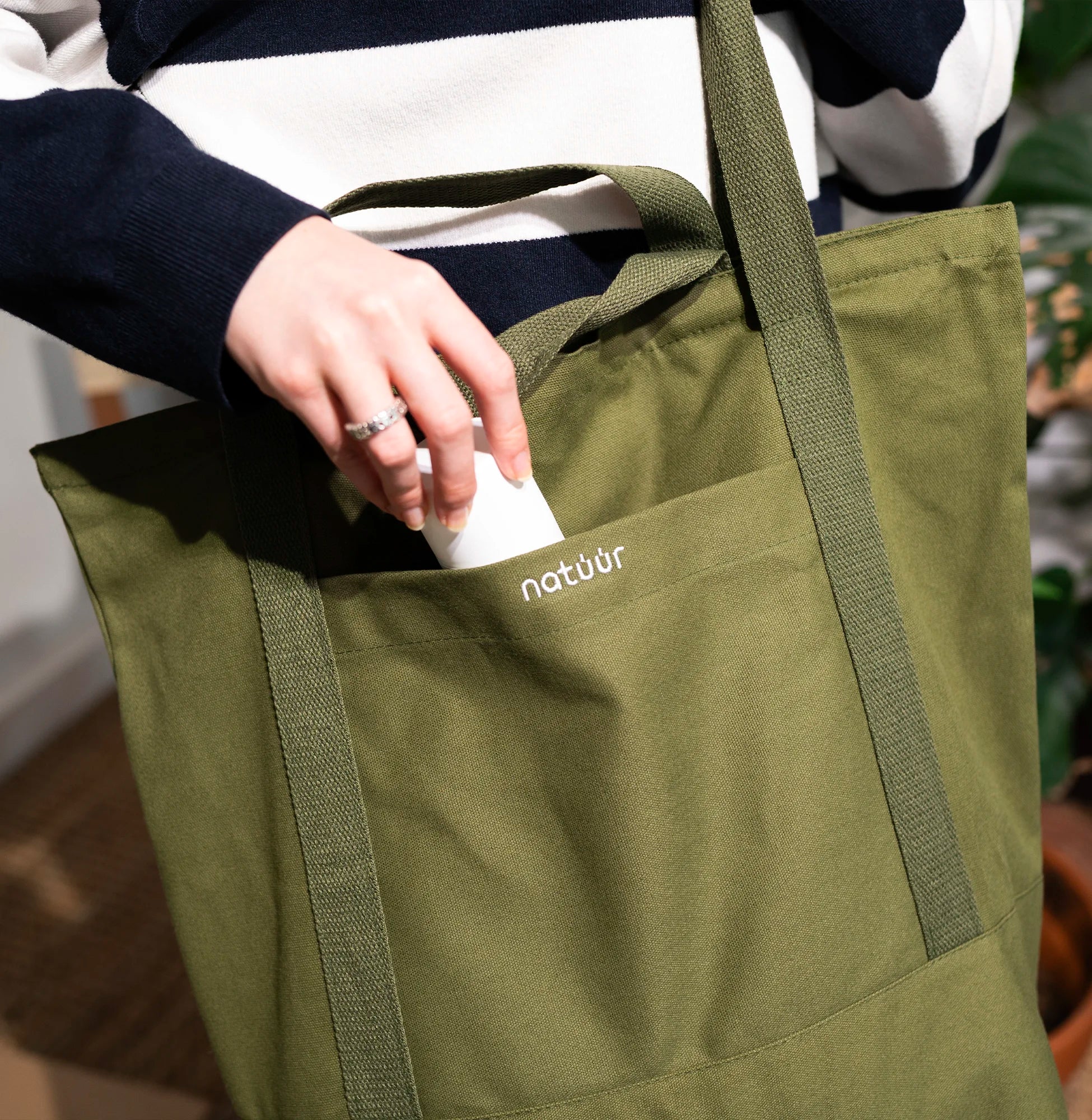 Person legt ein kleines Produkt in die Außentasche einer grünen Natuur-Tragetasche, ideal für umweltfreundliches Tragen und Organisation von Alltagsgegenständen.