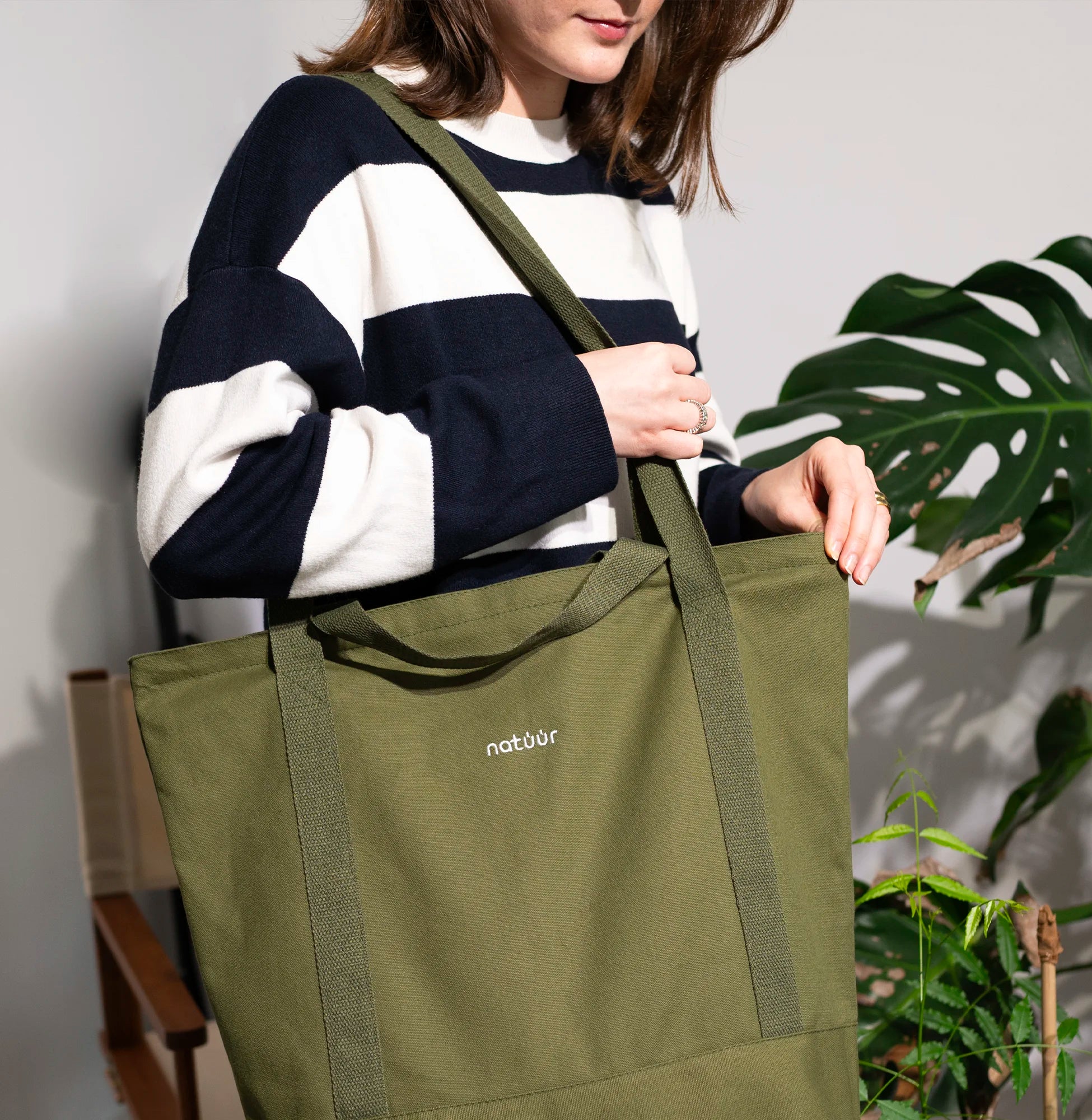 Person trägt eine grüne Natuur-Tragetasche über der Schulter, während sie einen gestreiften Pullover trägt, betont stilvolles und nachhaltiges Design für den Alltag, vor einer kulissenhaften Pflanzenlandschaft.
