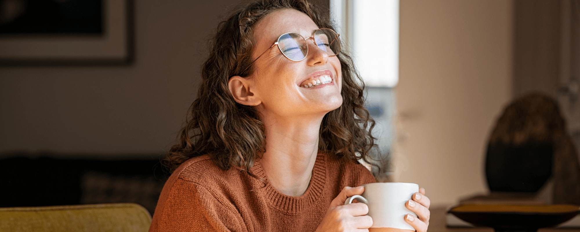 Eine Frau mit braunen Haaren hält eine Tasse in den Händen und lacht.