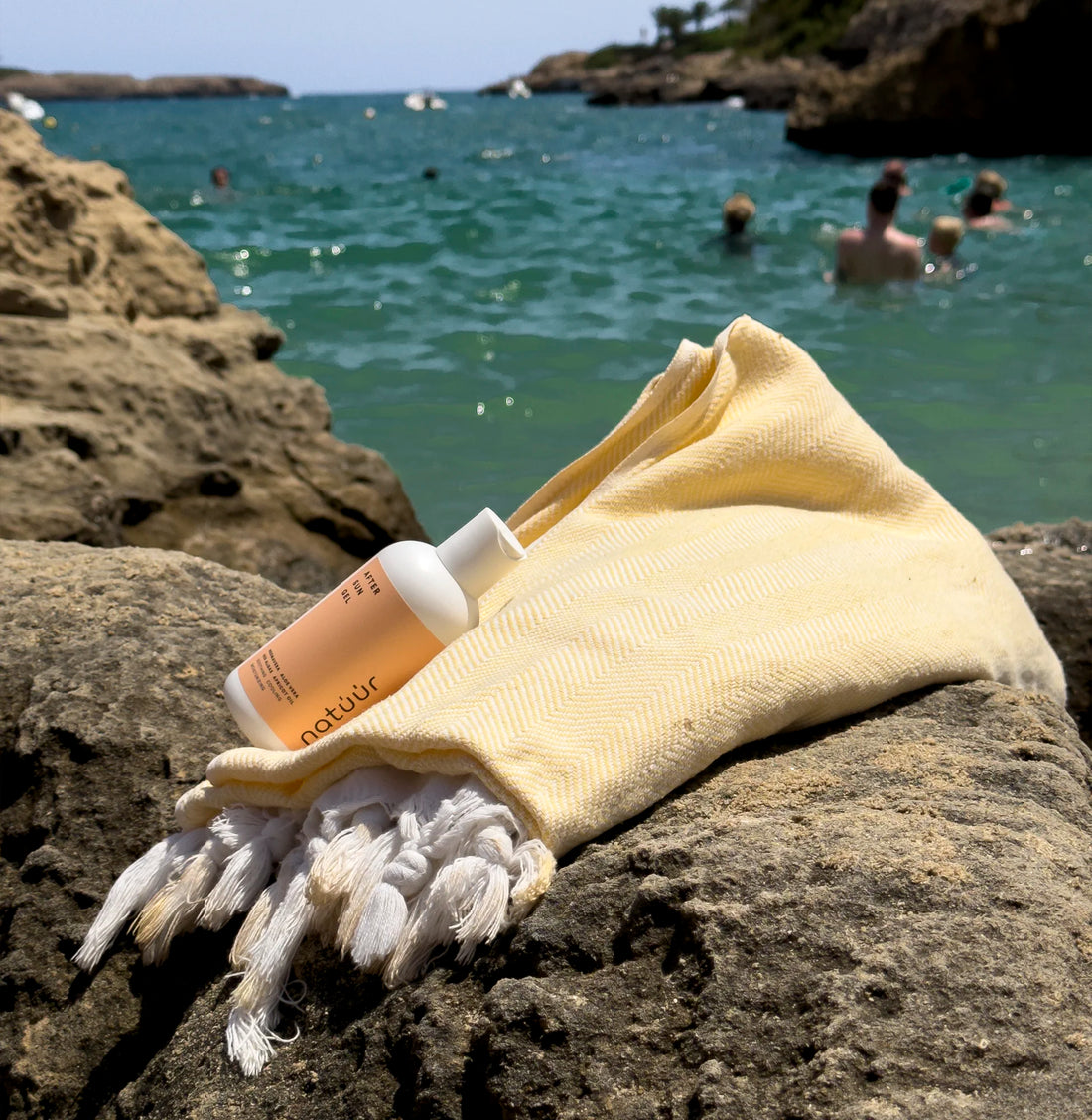 Man sieht das After Sun Gel der Marke natüür auf einem Stein, im Hintergrund das blaue Meer. Das Label auf der weißen Plastikflasche ist pfirsichfarben. Neben dem Produkt liegt ein Handtuch.