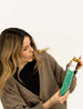 Eine Frau mit langen braunen Haaren und einer braunen Jacke hat die grüne Umverpackung des Scalp Tonics der Marke natüür in der Hand. Mit der anderen Hand zieht sie die Flasche mit dem Scalp Tonic raus.