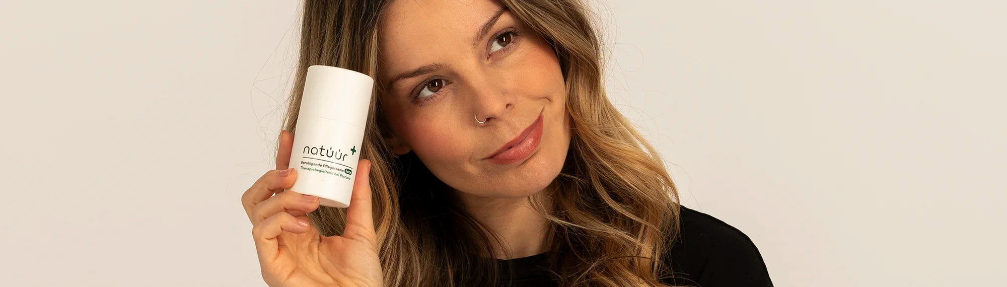 Eine Frau mit einem Lächeln auf den Lippen schaut Seitlich nach oben. Sie hält in ihrer Hand ein Hautpflege Produkt, die natüür Cream forte. 
