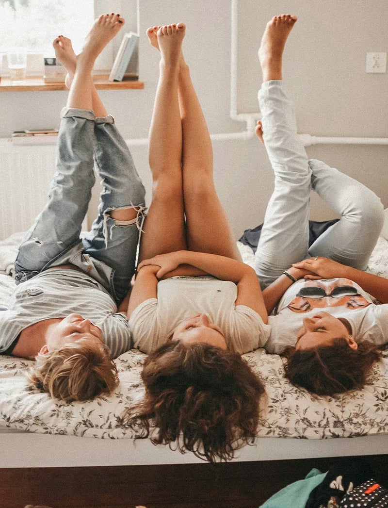 Man sieht drei Frauen mit langen Haaren, welche nebeneinander auf einem Bett liegen. Ihre Beine strecken sie in die Luft. 