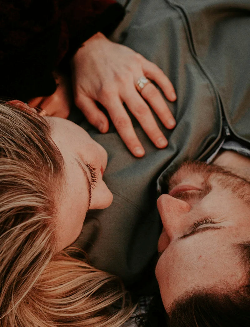 Ein Mann mit dunklem Bart und eine Frau mit blonden Haaren sind liegend zu sehen in einer Nahaufnahme. Sie hat ihren Kopf auf seiner Brust und die Hand auf seinem Oberkörper. Sie schauen sich an und wirken glücklich.