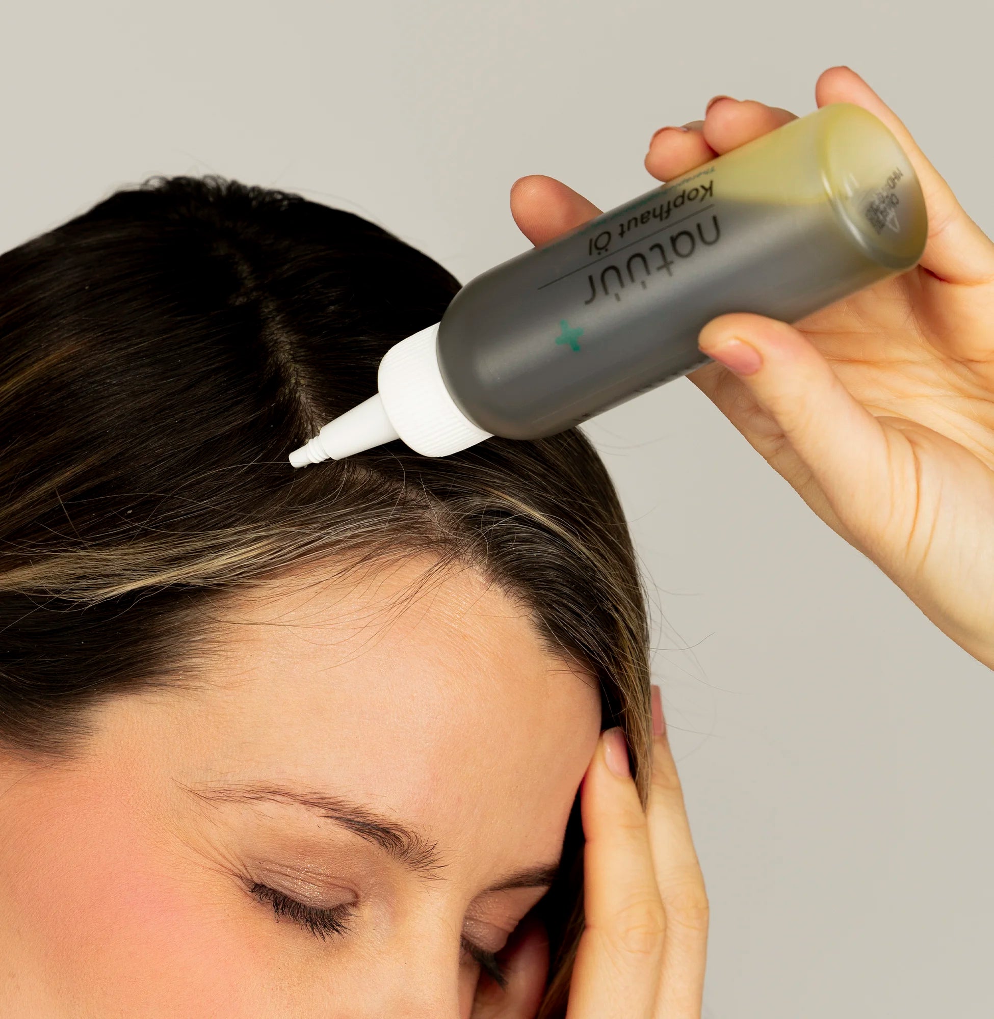 Eine Frau hält mit der Hand das Kopfhaut-Öl der Marke natüür in der Hand und setzt es an Ihre Kopfhaut an. Es handelt sich um ein Close-Up, das Gesicht ist etwas abgeschnitten.