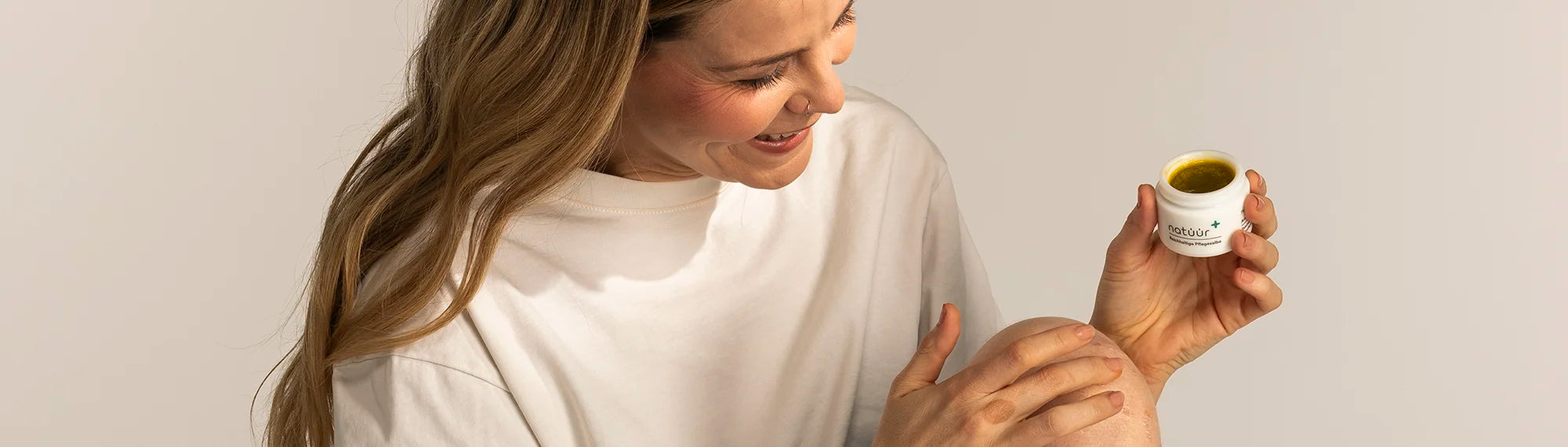 Eine Frau mit blonden Haaren im weißen T-Shirt gibt sich etwas natüür Pflegesalbe mit dem Finger auf ihr von Psoriasis betroffene Knie. In der anderen Hand hält sie das Tiegel. 
