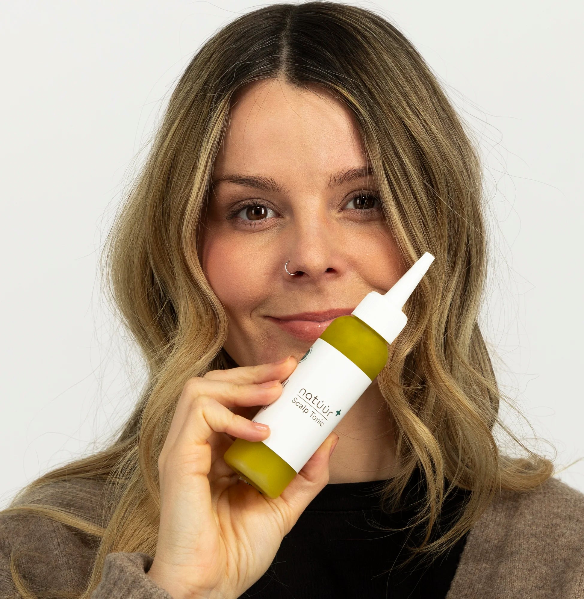 Eine glückliche Frau mit langen, blonden Haaren schaut in die Kamera. Ihre Lippen sind zu einem Lächeln angezogen. In der Hand hält sie das Scalp Tonic der Marke natüür, welches für die Kopfhaut Pflege von zB Psoriasis entwickelt worden ist. 