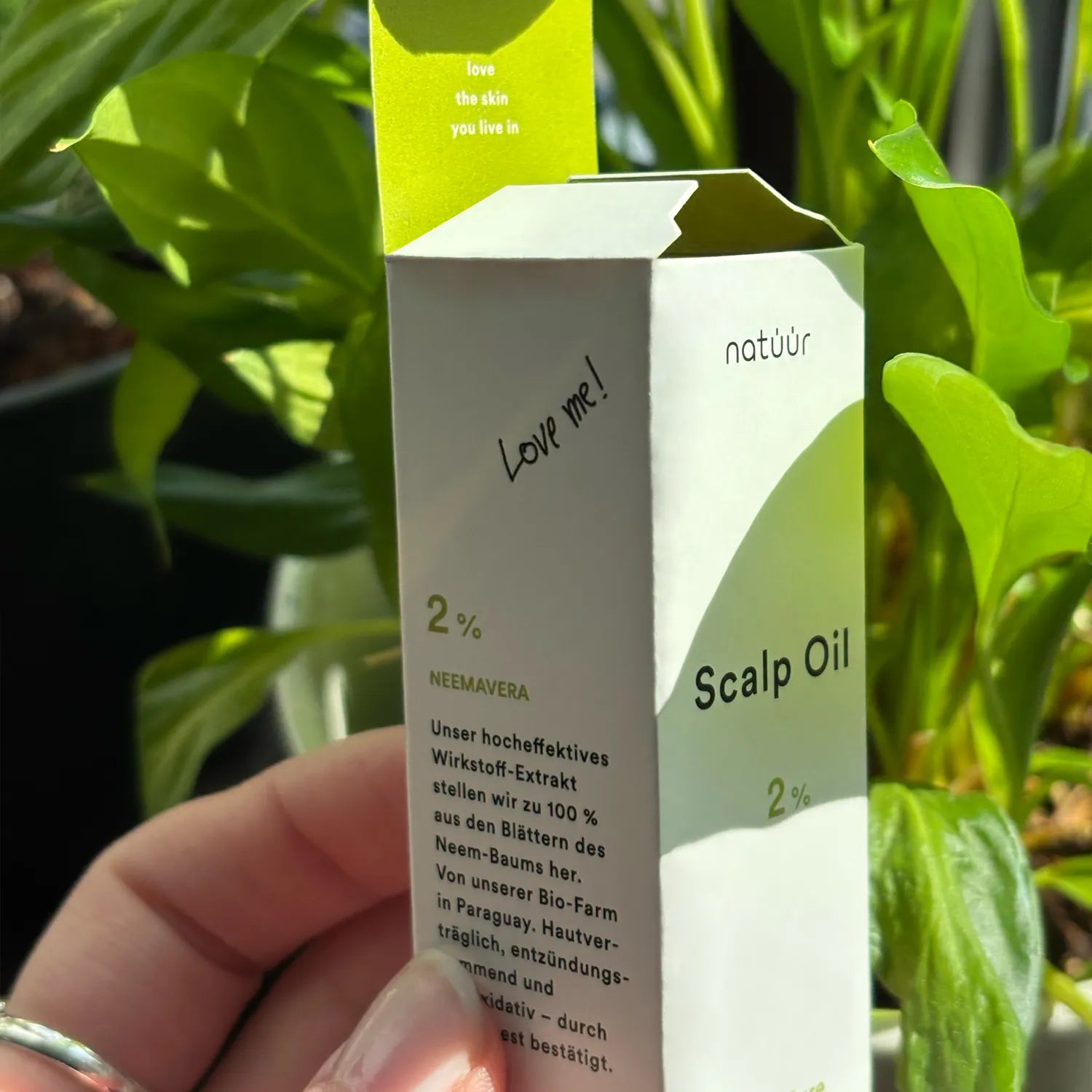 Naheaufnahme einer Schachtel Natuur Scalp Oil mit Neem-Extrakt, gehalten von einer Hand vor einem üppigen grünen Pflanzenhintergrund, mit Betonung auf natürliche Hautpflege und den Texten 'Love me!' und 'Love the skin you live in.