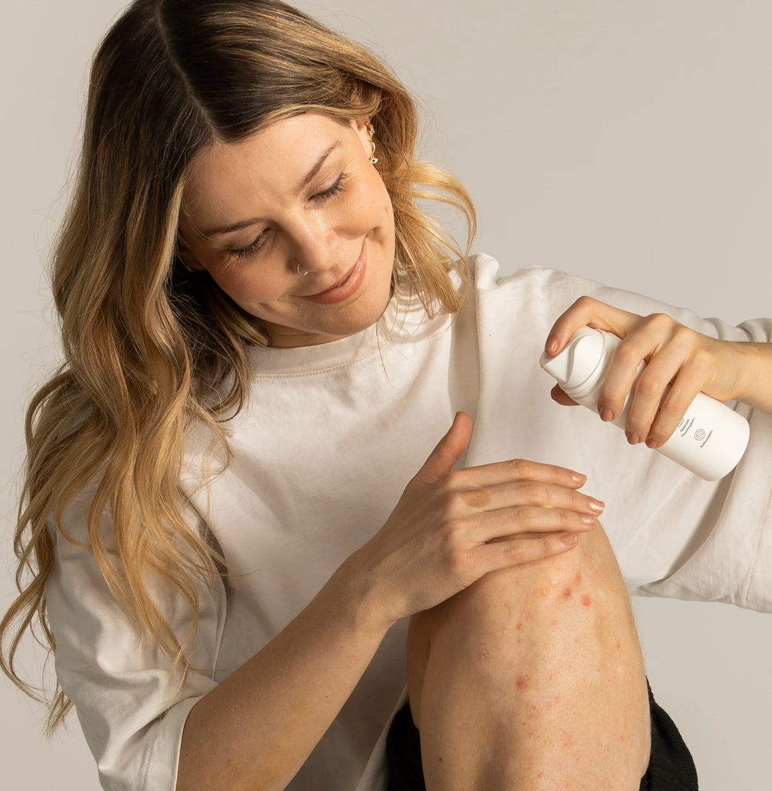 Das Gesicht der Frau schaut nach unten, denn sie sitzt auf dem Boden. Ihr Bein ist angewinkelt, sodass ihr Knie gut zu erkennen ist. Dieses ist von Psoriasis (Schuppenflechte) betroffen. In der einen Hand hält sie die natüür Pflegecreme, mit der anderen cremt sie sich ihr Knie ein.